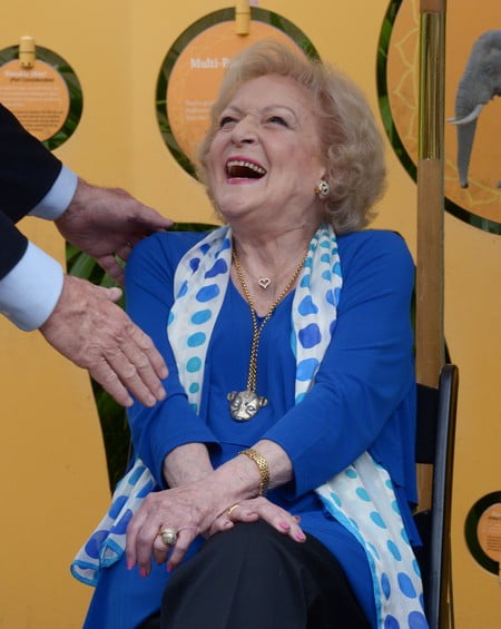 Betty White is dressed in a bright royal blue dress coat with a dainty white dress scarf with blue polka dots draped around her neck. Her hands are placed on her lap and a man who is out of shot has his hands on her elbow and shoulder. Betty White is looking to this man and laughing happily.