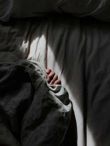 A curled hand peaking out from under gray bed sheets in a beam of sunlight shaped in a rectangular pattern. The surrounding bed sheets are dark.
