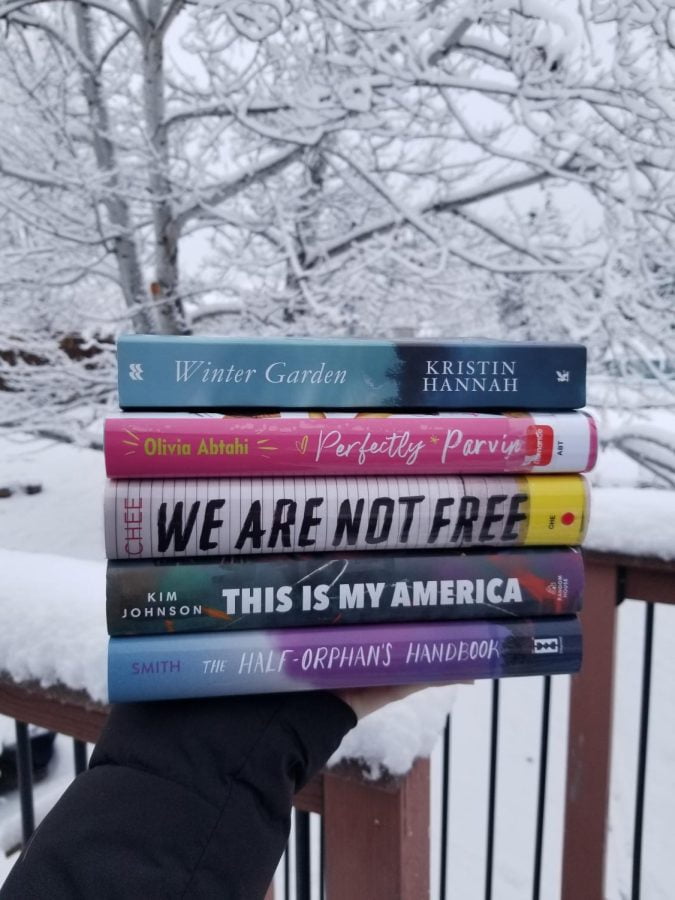 In the picture, a hand holding a stack of five books, and the backdrop is a snow covered tree. The books featured from top to bottom are: Winter Garden, Perfectly Parvin, We Are Not Free, This Is My America, and The Half-Orphans Handbook.