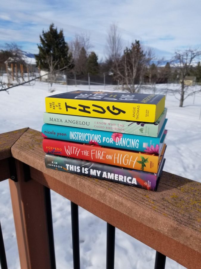 This image shows a stack of books on a wooden railing. The background is a snow-covered landscape. The stack begins with The Hate U Give on top and then in descending order: I Know Why the Caged Bird Sings, Instructions for Dancing, With the Fire On High, and finally This is My America.