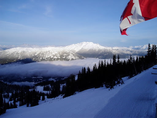 The image shows a snow covered mountain in the distance with a baby blue colored sky above. Silhouetted trees are in the fore ground. A Canadian flag flies in the upper right corner.