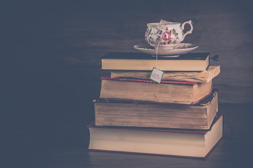 Image depicts a stack of old books with their pages facing outwards. On top there is a tea cup on a saucer with a pink floral pattern and a tea bag string hanging out of it.