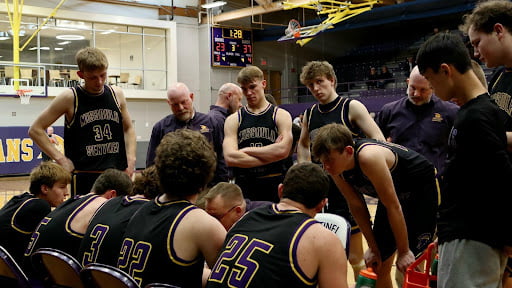This image shows the boys Varsity basketball team for Sentinel. Three bald men stand in the background. The head Coach, is barely visible in the center of the image because he is crouched down in the ring of high school boys giving directions. All of the players are dressed in their black jerseys with purple numbering.
