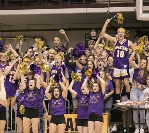 The image shows students on bleachers cheering and waving pom-poms in their purple Golden Goat shirts.