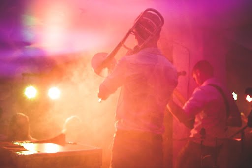 Image shows a guy playing a trombone in the center and a guitarist to the right. They are both wearing what seems to be white button down shirts. The lights are casting pink, orange and light yellow colors onto them.