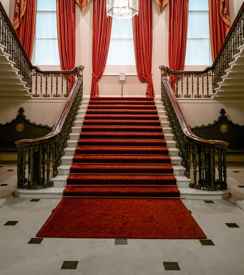 This image shows a red carpeted stair case with wooden grand railings.