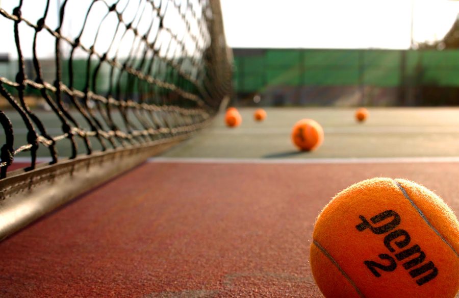 This image shows a angled photograph of a red and green tennis court. There is an orange tennis ball in the lower right corner with several other balls in the background. The net is on the left.