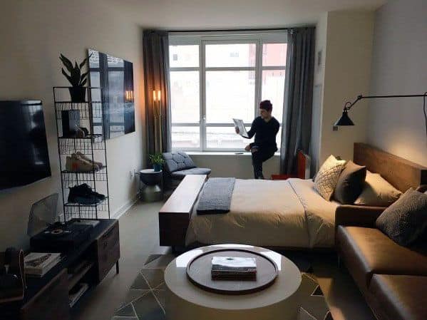 Image of a clean small hotel like room with a large window and a man standing at it.