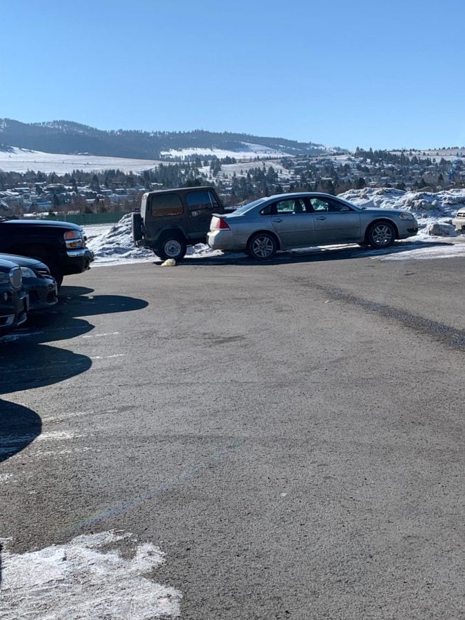 This image shows a high school parking lot in the winter. A little silver car in front of a jeep are in the center of this picture with snow around and snowy mountains in the background.