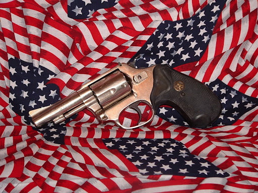 This picture shows many small American flags positioned as a backdrop with a small pistol placed over the top. The gun has a coppery metal look to the barrel, muzzle and cylinder. The handle grip is a dark brown.