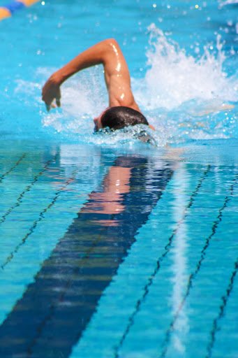 This image shows a man swimming through vibrant light blue water at a pool. His arm is arched up out of the water, he is wearing a black swim cap.