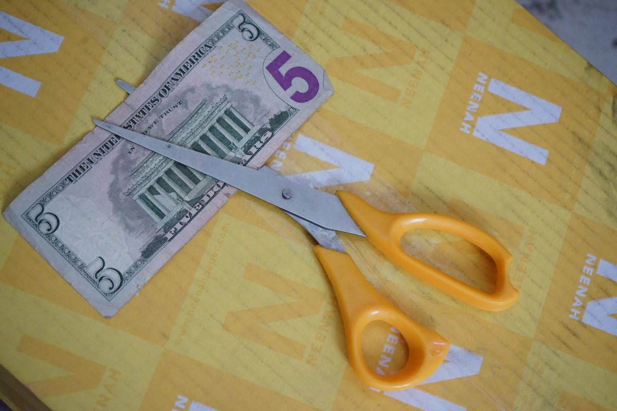 A pair of scissors posed like they are cutting a five dollar bill